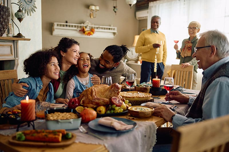 A multi-generational family enjoys a festive meal together around a dining table with a roast turkey centerpiece, various dishes, and lit candles, conveying a sense of joy and togetherness.
