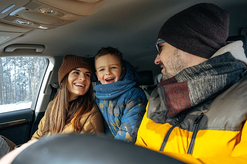 A family of three, with a smiling woman and a laughing child sitting in the backseat of a car, and a man in the driver's seat turned towards them, all dressed in warm winter clothing.